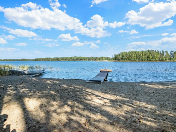 A lakeside beach with a boat docked, a wooden deck, and a forest in the background.
