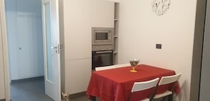 Una zona de menjador reduïda amb una taula vermella i cadires blanques, un microones i un forn al fons.