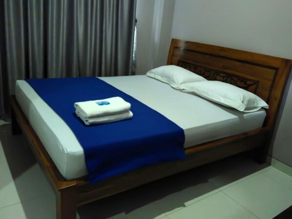 A neatly made bed with a blue bedspread and white pillows, a wooden headboard, and a white wall in the background.