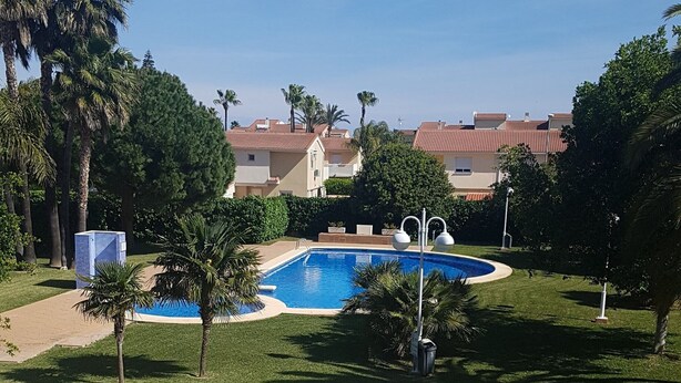 Un quartier résidentiel doté d’une piscine entourée de palmiers, avec un ciel bleu dégagé.
