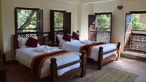 A room with two wooden beds, each with white bedding and red pillows. There are windows with wooden shutters, and a balcony with a decorative railing.