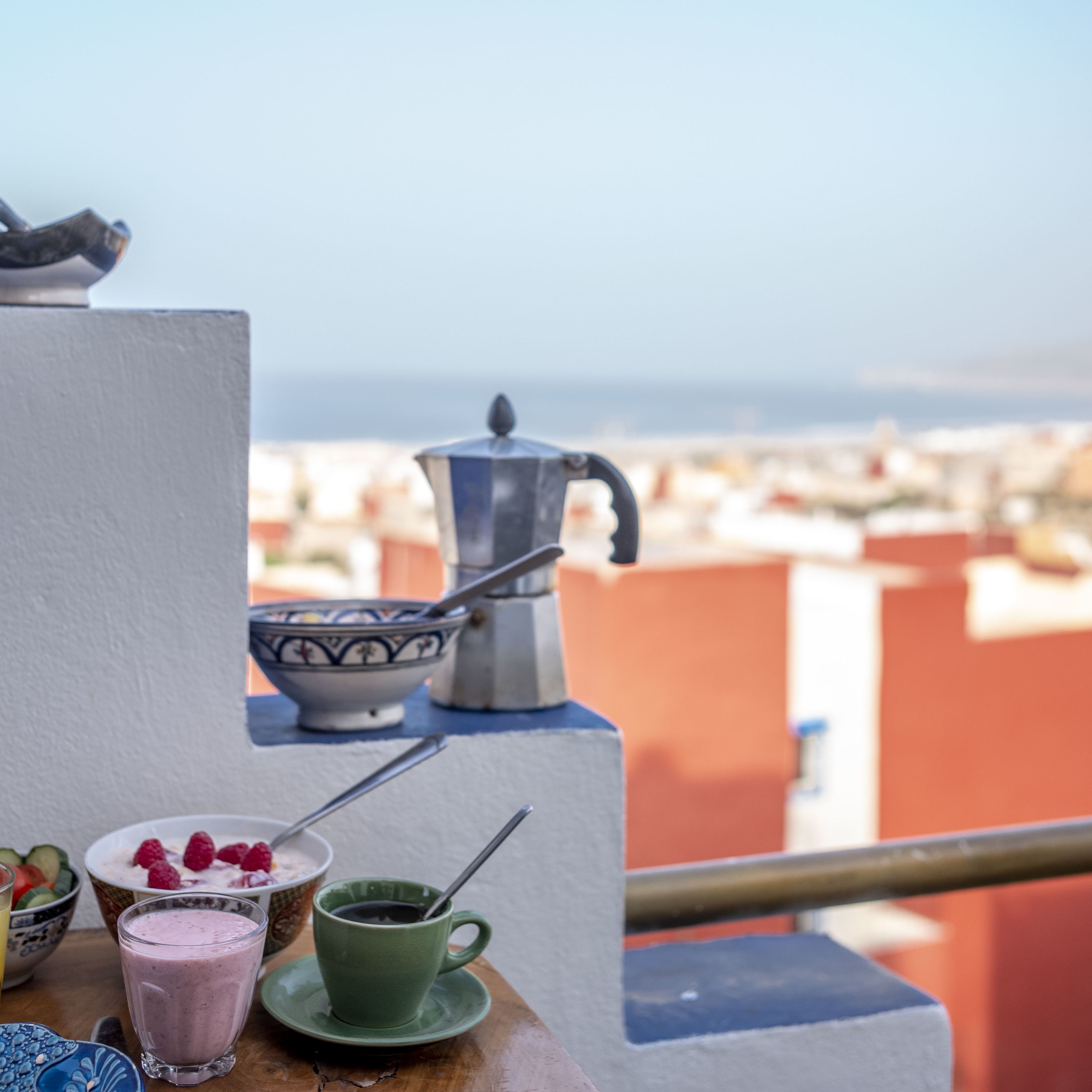 A balcony with a view of the city, a coffee pot, and various food items on a table.