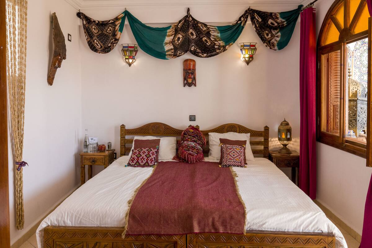 Una cama con cabecera de madera, dos mesitas de noche, almohadas estampadas, una colcha roja y adornos colgantes en la pared.
