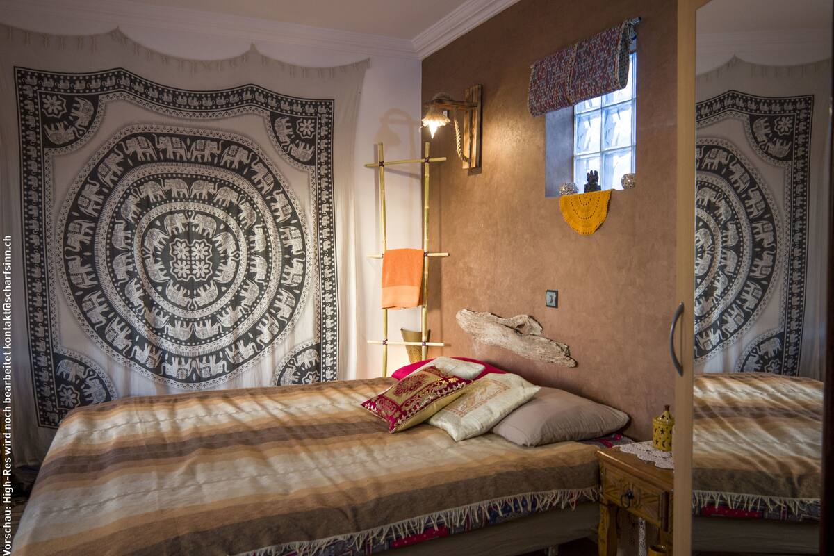 Un dormitorio con una cama grande, una mesita de noche, un tapiz en la pared y una ventana con cortinas.