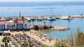 A coastal marina with boats docked, a sandy beach with umbrellas, and a building with a red roof. - Fantastic 2 bed 2 bath beachside penthouse with pool and parking (Marbella)