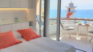 A modern bedroom with a large bed, two pillows, a glass table with a clock, and a view of the sea and a lighthouse. - Fantastic 2 bed 2 bath beachside penthouse with pool and parking (Marbella)