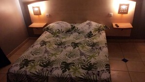 Een bed met een tropisch bladmotief dekbed, twee nachtkastjes met lampen, en een betegelde vloer.