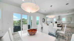 A modern kitchen with white cabinetry, a dining area with a white table and chairs, and a large window overlooking a balcony. - 30A Retreat - 2 Homes, Gulf Views, Great for Large Groups! (Santa Rosa Beach)