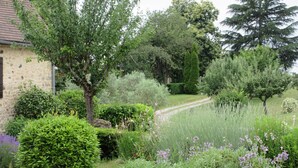 A lush garden with various plants and trees, a stone building, and a pathway. - Domaine de l'Homme: The Red House with swimming pool (ST VINCENT LE PALUEL)