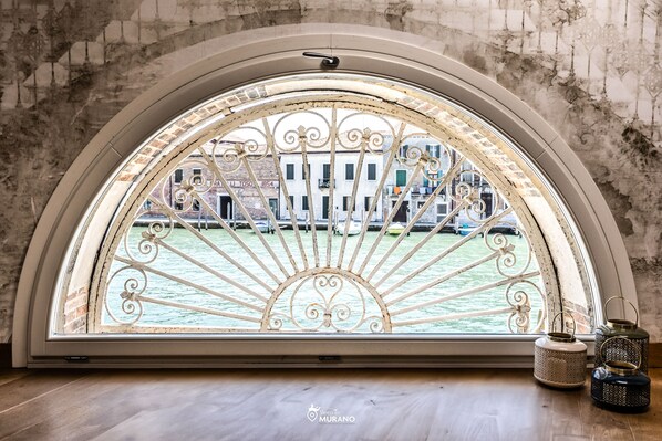 A room with a large arched window overlooking a canal with boats, a stone wall, and a view of buildings.