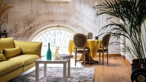 Een kamer met een gele bank, een klein tafeltje met een vaas en boek, een eettafel met stoelen en een grote plant.
