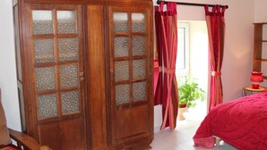 A room with a wooden wardrobe, a bed with a red quilt, a small table with a lamp, and a window with curtains.