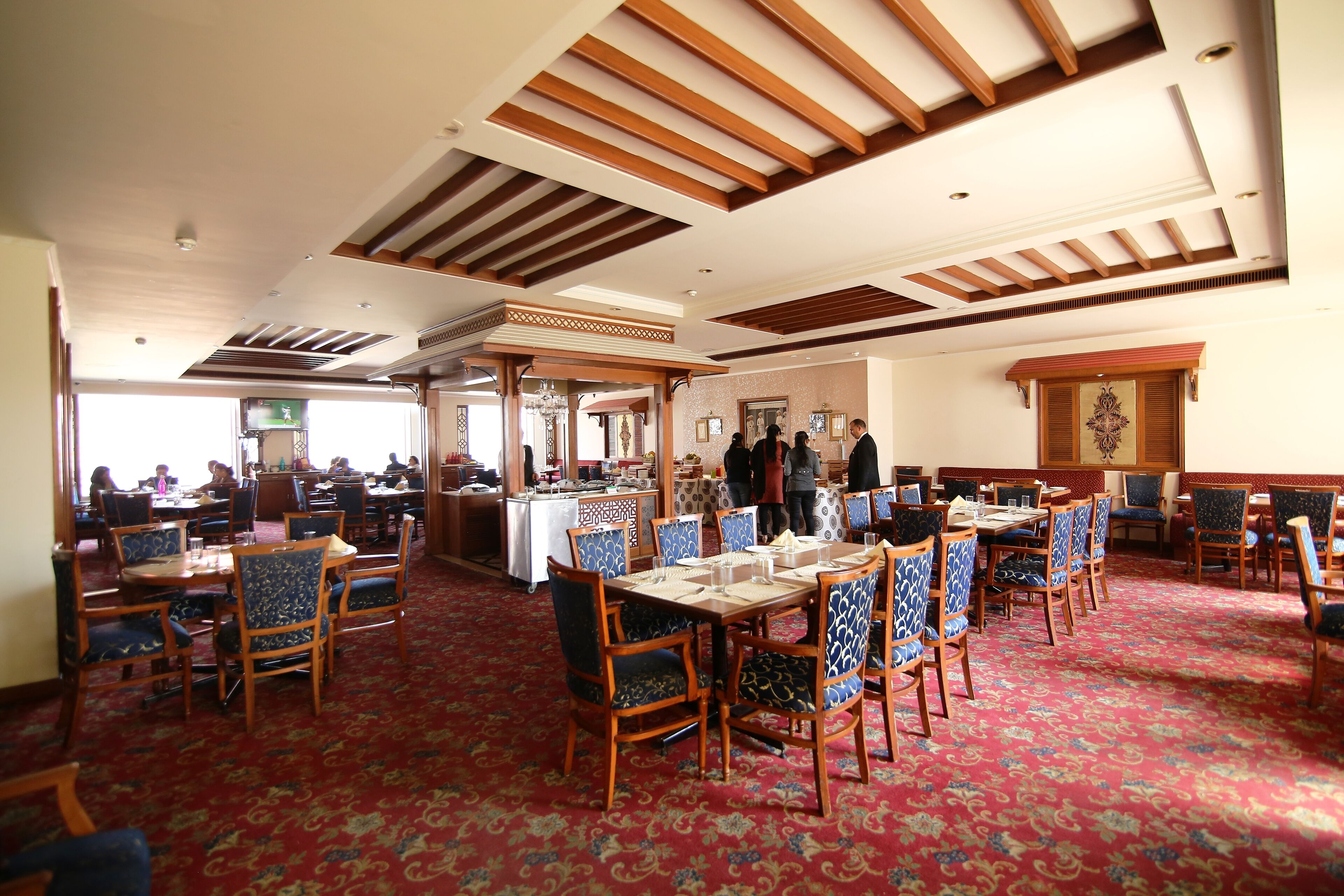 A spacious dining area with multiple tables and chairs, a patterned carpet, and a high ceiling with wooden beams.