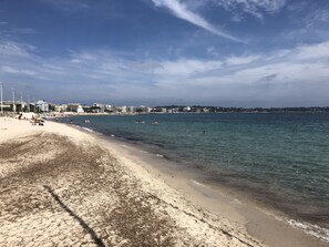 Uma praia com águas cristalinas, areal arenoso e edifícios ao fundo.