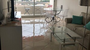 A modern living room with a glass coffee table, a sofa with a teal pillow, and a balcony with a red awning. - 3 rooms apartment renovated with panoramic sea view (antibes)