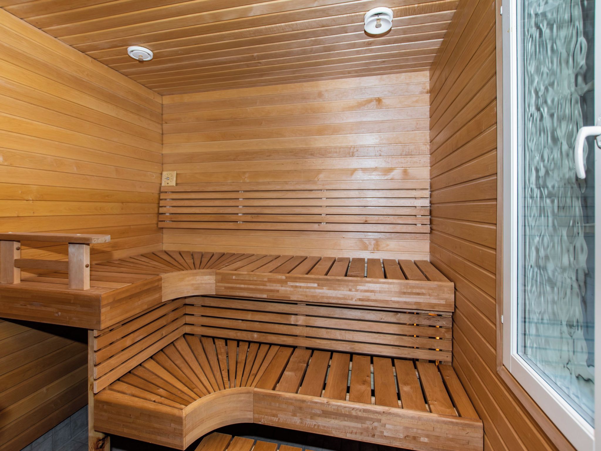 A sauna with wooden benches arranged in a semi-circular pattern, a glass door, and a ceiling with recessed lighting.