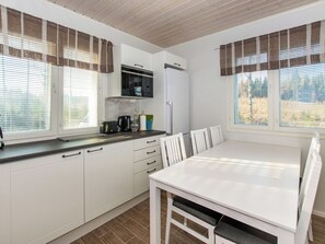 Una cocina moderna con armarios blancos, un microondas negro y un refrigerador blanco. Hay una mesa de comedor con sillas y una ventana con persianas.
