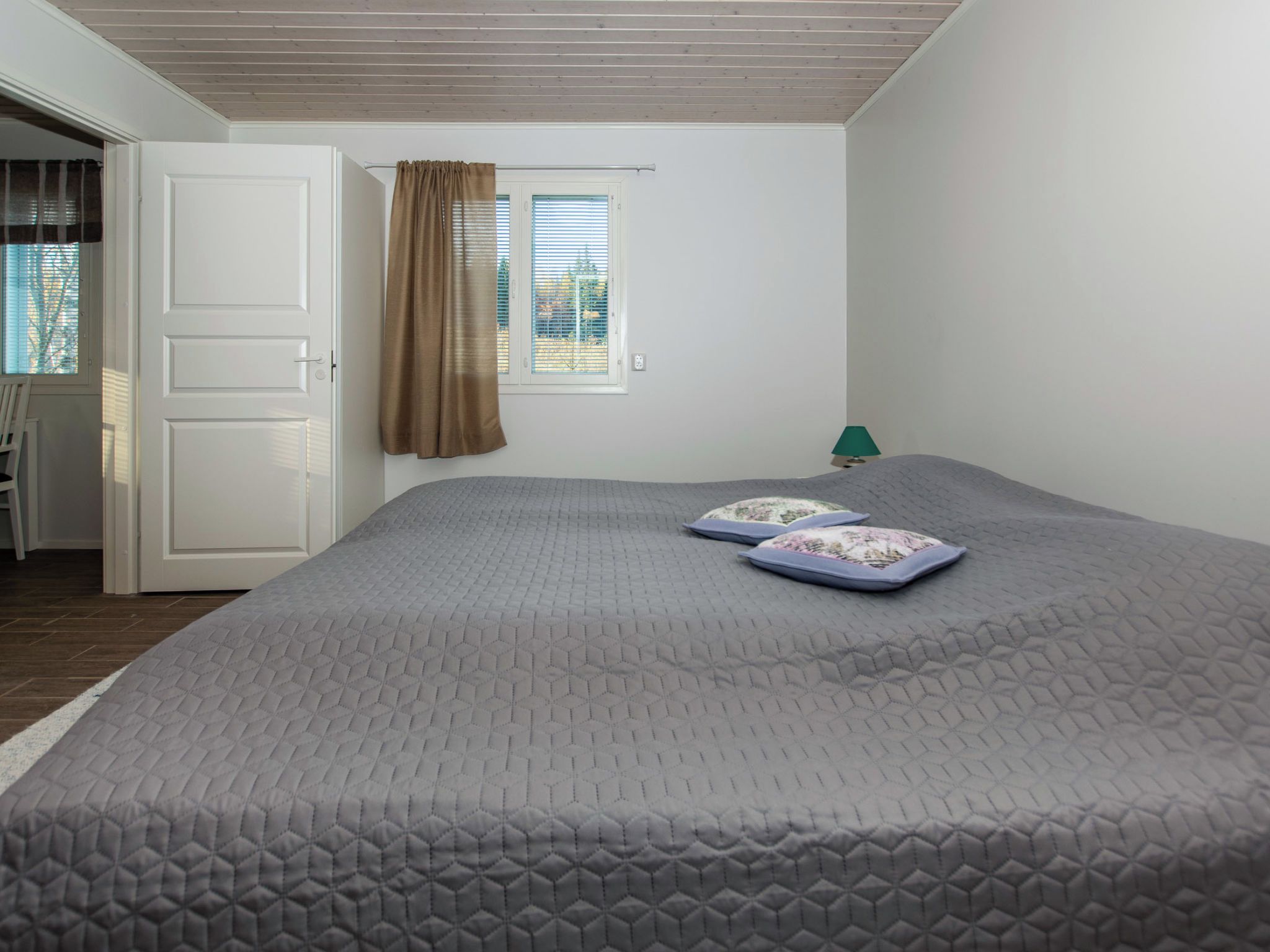 Une chambre avec un grand lit, une fenêtre avec des stores, une porte et une petite table avec une lampe.