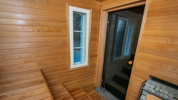 A wooden sauna room with a bench, a window, and a door leading to another room.