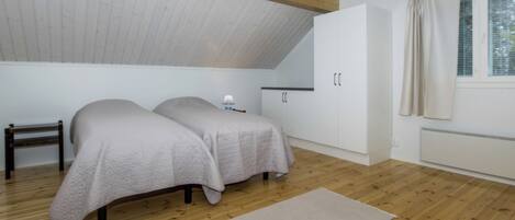 Ein Schlafzimmer mit schrägem Dach, Holzboden, zwei Betten mit weißer Bettwäsche, einem Nachttisch, einer Lampe, einem Kleiderschrank und einem Fenster mit Vorhängen.