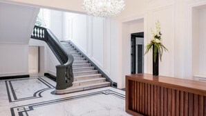 A grand lobby with a marble floor, a staircase with black balusters, a wooden reception desk, and a chandelier.