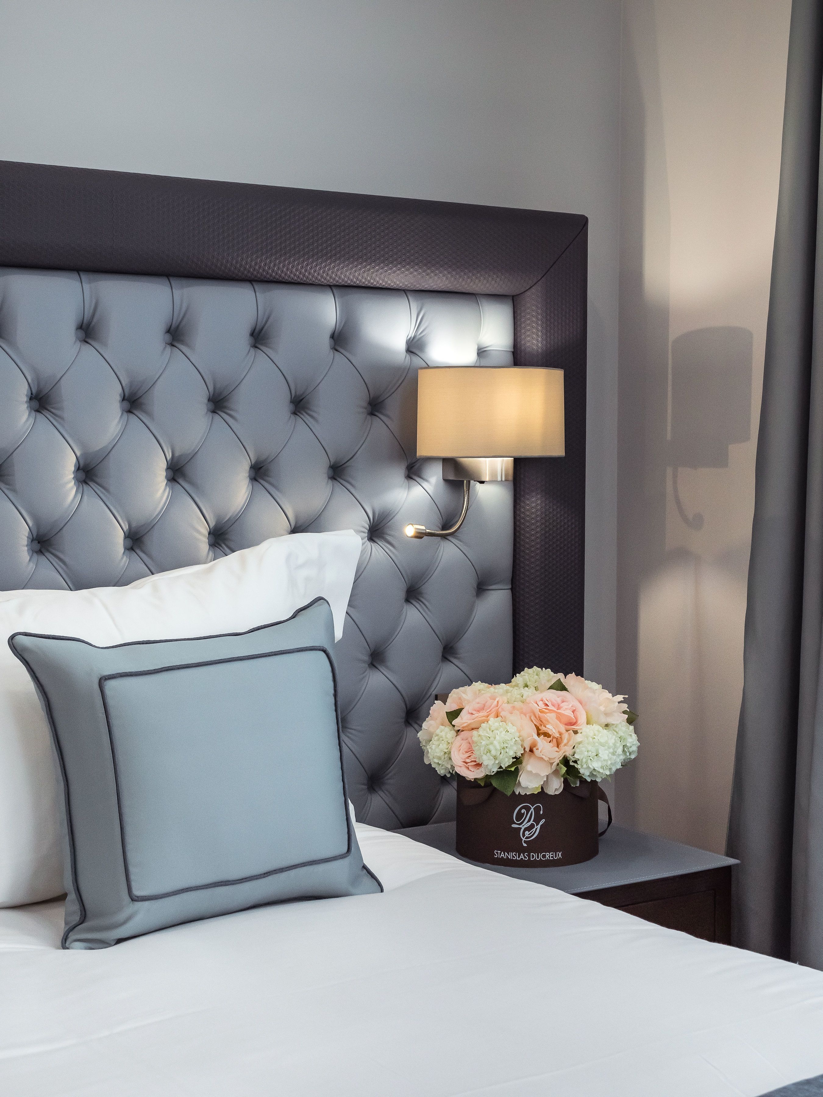Een net opgemaakt bed met een gestoffeerd hoofdeinde, een bedlampje, een vaas met bloemen en een nachtkastje.