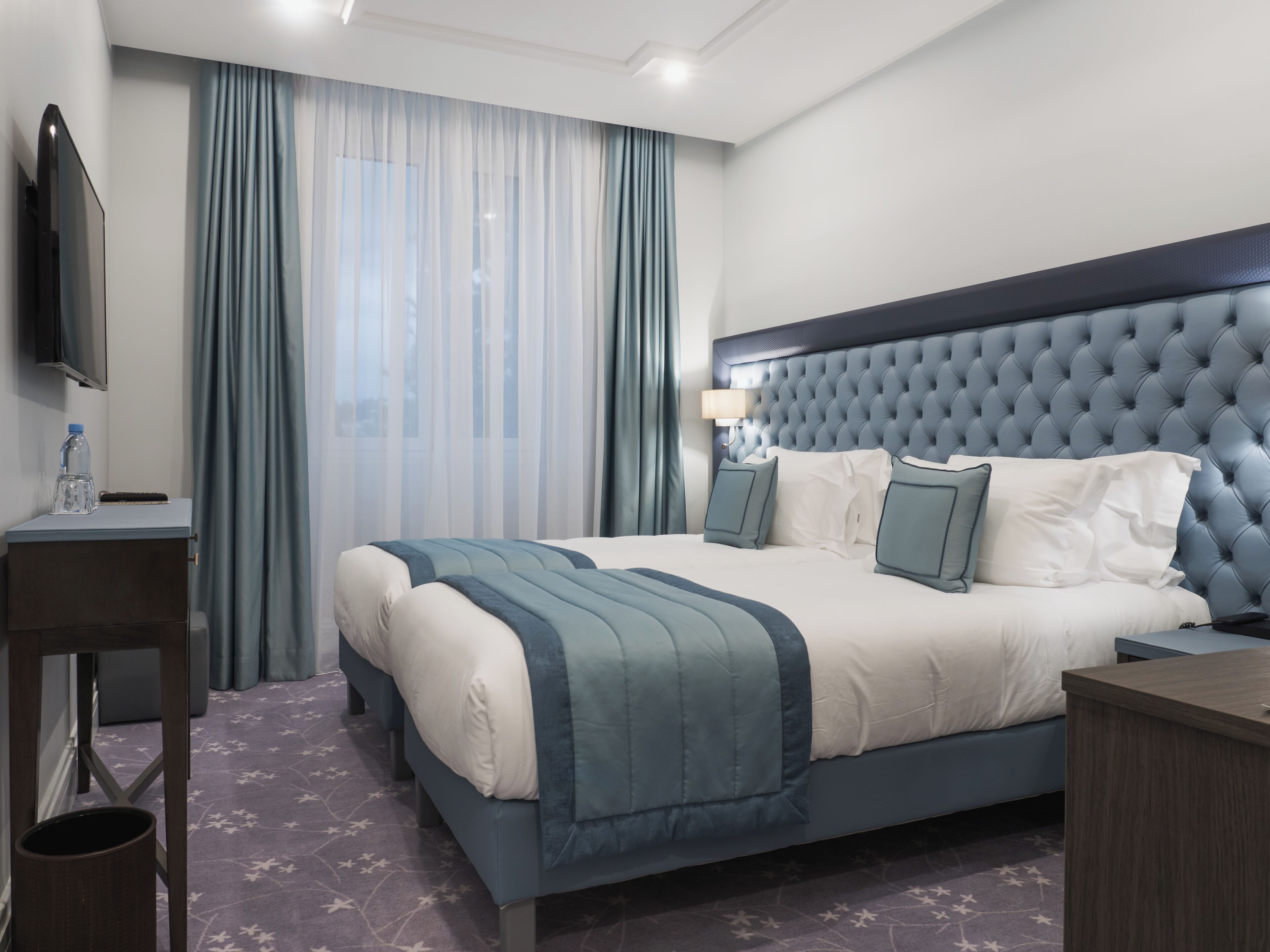 A hotel room with a bed, a TV, a bedside table, a lamp, and a window with curtains.