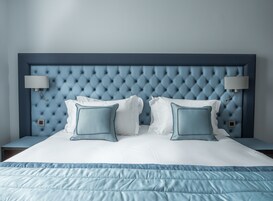 Una cama bien hecha con cabecera capitonada, dos lámparas de noche y una almohada decorativa azul.