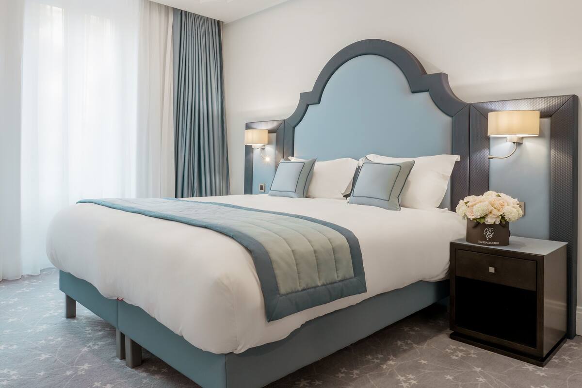 Une chambre d’hôtel avec un grand lit, deux tables de chevet, une petite table avec un vase de fleurs et une fenêtre avec des rideaux.