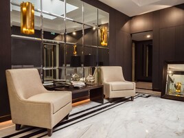 Un lobby moderno con dos sillones color beige, una mesa de centro de vidrio y un espejo grande que refleja la estancia.