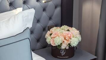 Un lit bien fait, avec du linge de lit blanc et un oreiller bleu clair, une tête de lit capitonnée, une table de chevet avec une lampe et un vase de fleurs.
