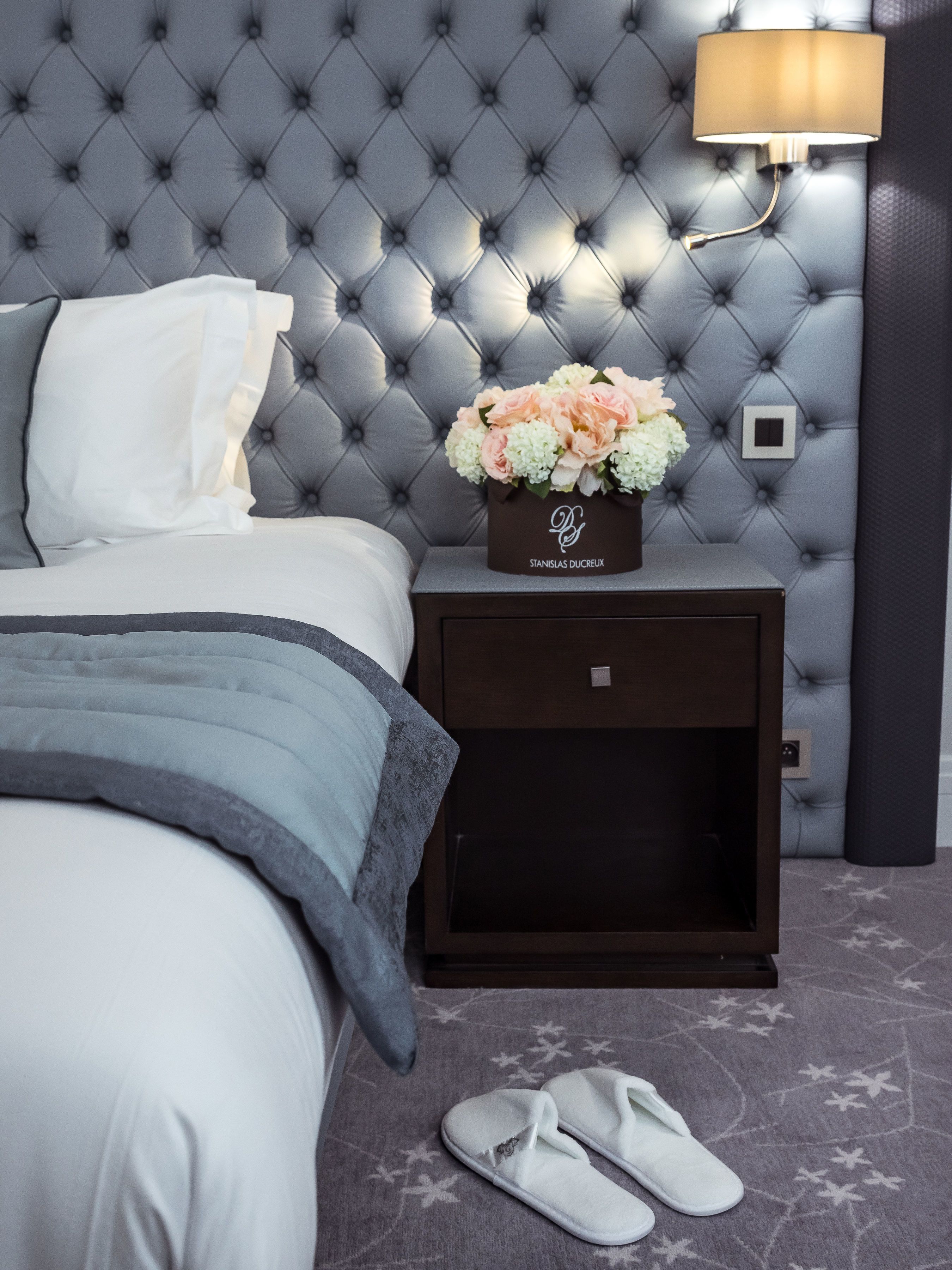 A bedroom with a tufted headboard, a bedside table with a lamp, a floral arrangement, and a pair of slippers on the floor.
