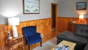 Un salon avec un fauteuil bleu, un canapé foncé, une table basse en bois et une lampe sur une table d’appoint.
