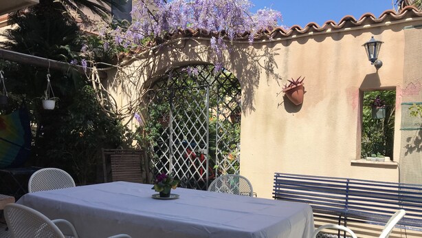 An outdoor patio with a table and chairs, surrounded by a pergola with purple flowers, a wall-mounted lantern, and a small window with plants. - Holiday home the fish house (Palermo)
