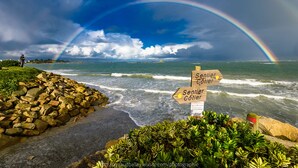 Une plage avec un arc-en-ciel, une côte rocheuse et un panneau indicateur.