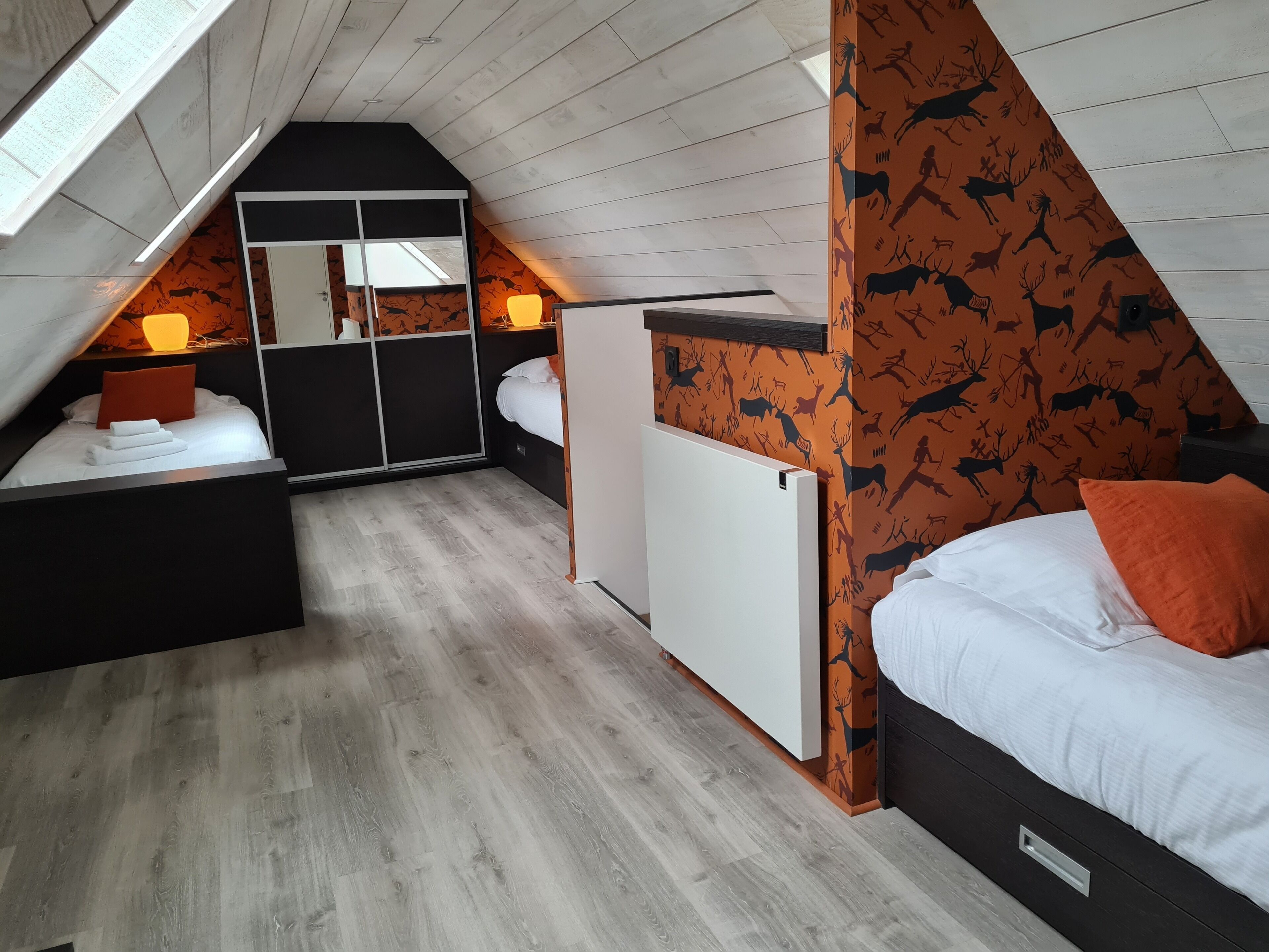 A bedroom with a sloped ceiling, a bed with white bedding and orange pillows, a bedside table with a lamp, a wardrobe, and a radiator.