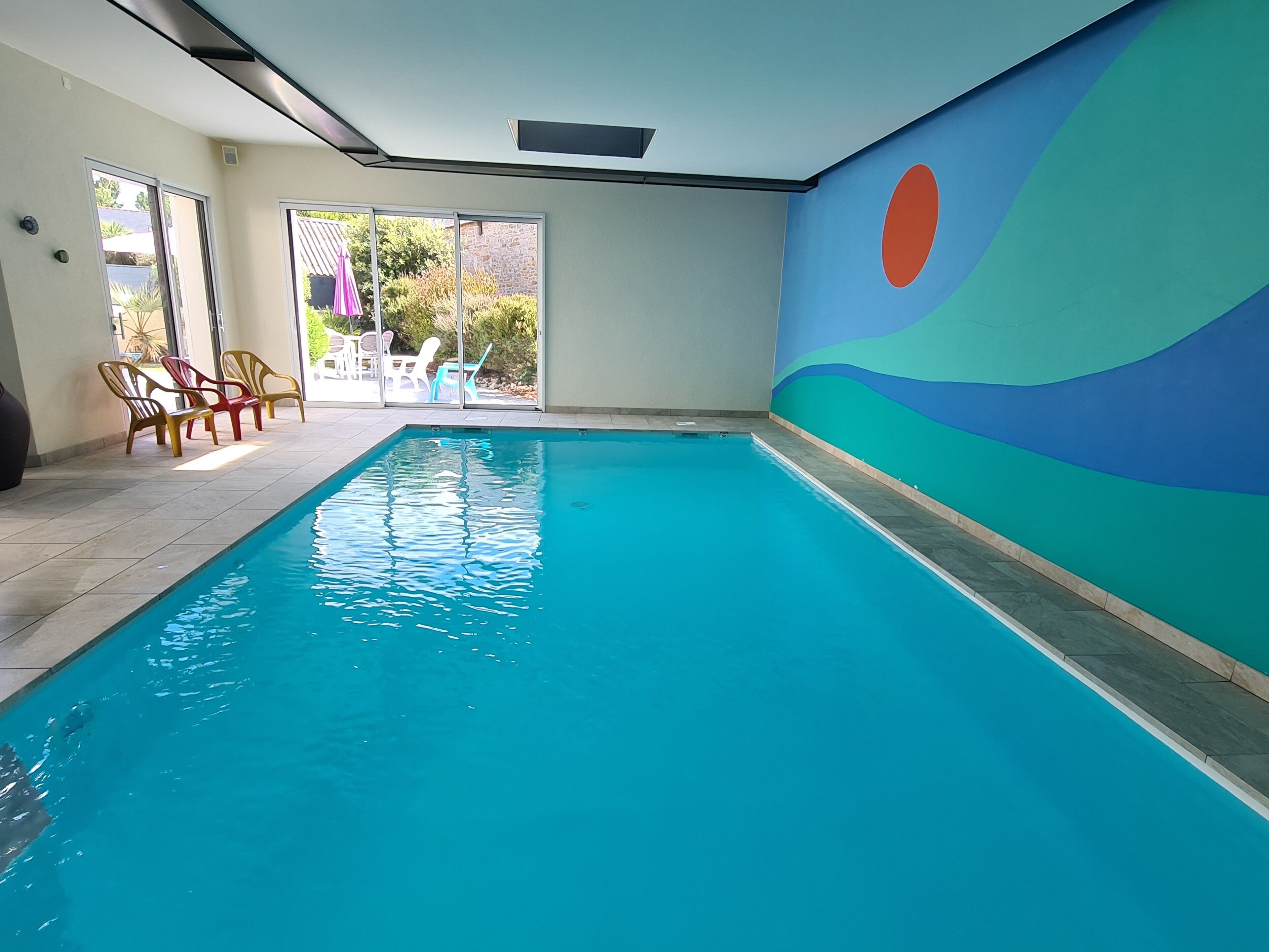 Une piscine intérieure à l’eau d’un bleu limpide, dotée d’un sol carrelé et offrant une vue sur le mobilier de patio et la verdure à travers des portes vitrées.