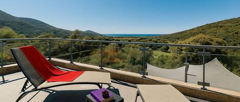 Un balcone con una sedia rossa, un tavolino con un libro e una vista panoramica su colline e un lago.