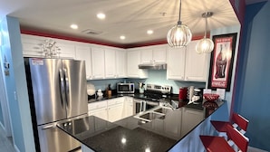 A modern kitchen with a central island, stainless steel appliances, and a bar area with red stools. - Gulf-Front Calypso 1BR/2BA | Free Beach Service (Panama City Beach)