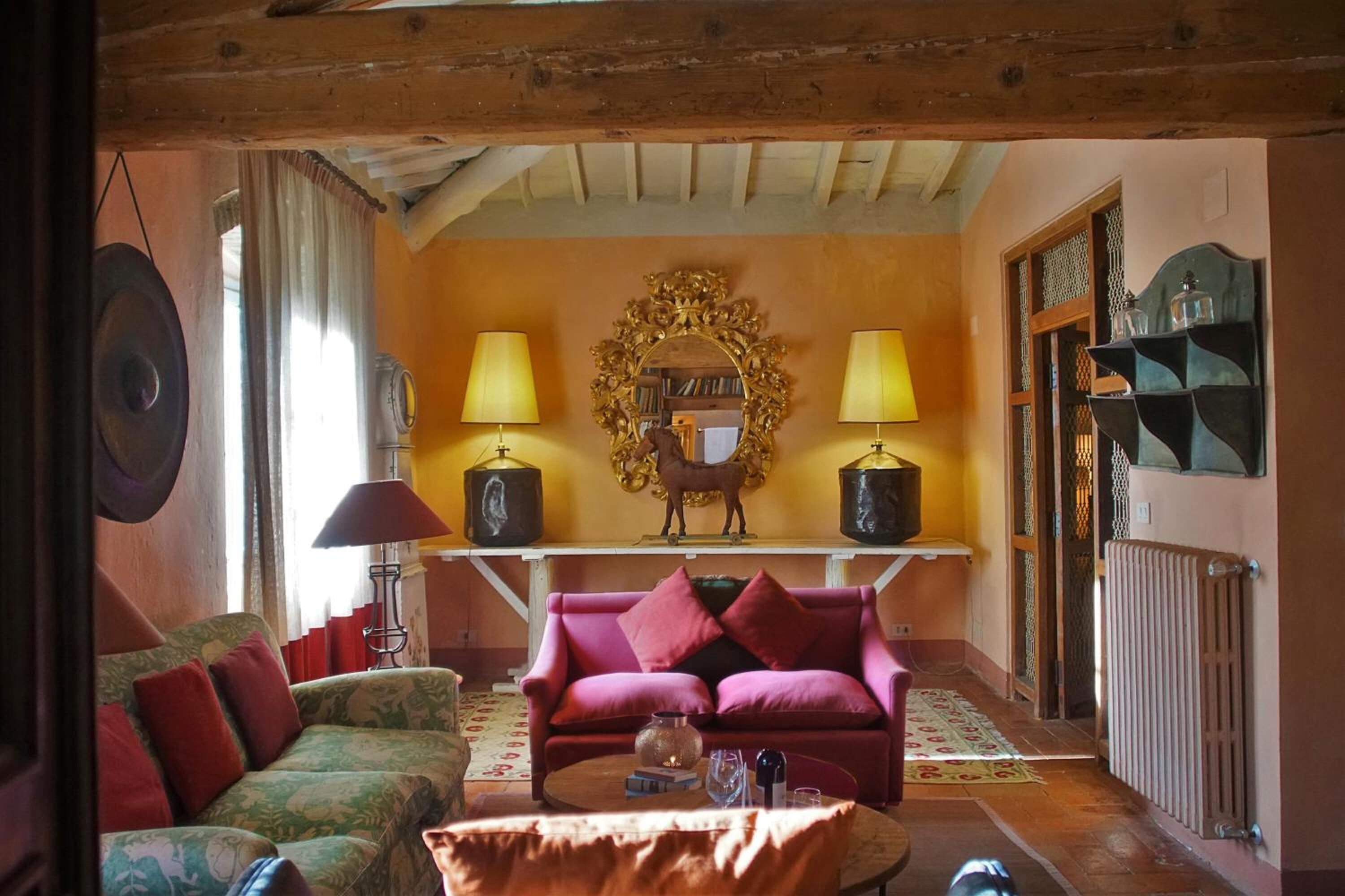 A cozy living room with a wooden ceiling, a sofa, a pink armchair, a coffee table with books and a glass, and a decorative mirror.
