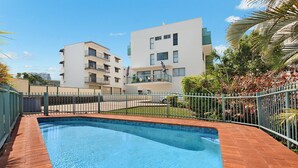 Ein Swimmingpool mit einer gefliesten Kante, umgeben von einem Pflasterweg. Im Hintergrund ein mehrstöckiges Gebäude mit Balkonen und einem verschlossenen Eingang.