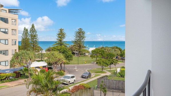 A residential area with a view of the ocean, featuring a road with parked cars, a white building, and various trees. - Fair Havens unit 4 Kings Beach QLD (KINGS BEACH)