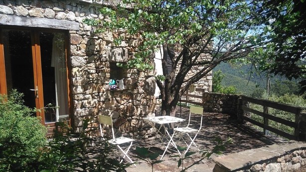 A stone building with a wooden door, a small table with chairs, and a tree in the background. - Gîte "La Terrasse". A real "Eagle's Nest" for two people. (Beaumont)