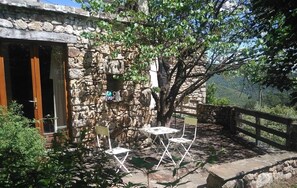 Un bâtiment en pierre avec une porte en bois, une petite table entourée de chaises et un arbre en arrière-plan.