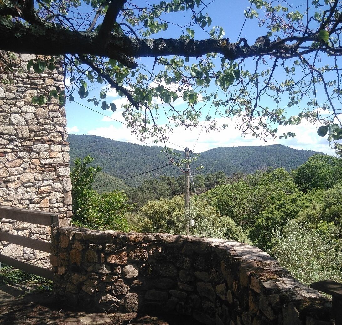 Mural de pedra com cerca de madeira, um poço de pedra e vista para uma cadeia montanhosa arborizada.