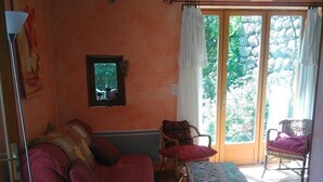 A living room with a red sofa, a wooden chair, a small table, and a view of a garden through a glass door. - Gîte "La Terrasse". A real "Eagle's Nest" for two people. (Beaumont)