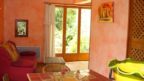 A room with a red sofa, a dining table with a mosaic tile top, a wicker chair, and a sliding glass door leading to an outdoor area. - Gîte "La Terrasse". A real "Eagle's Nest" for two people. (Beaumont)