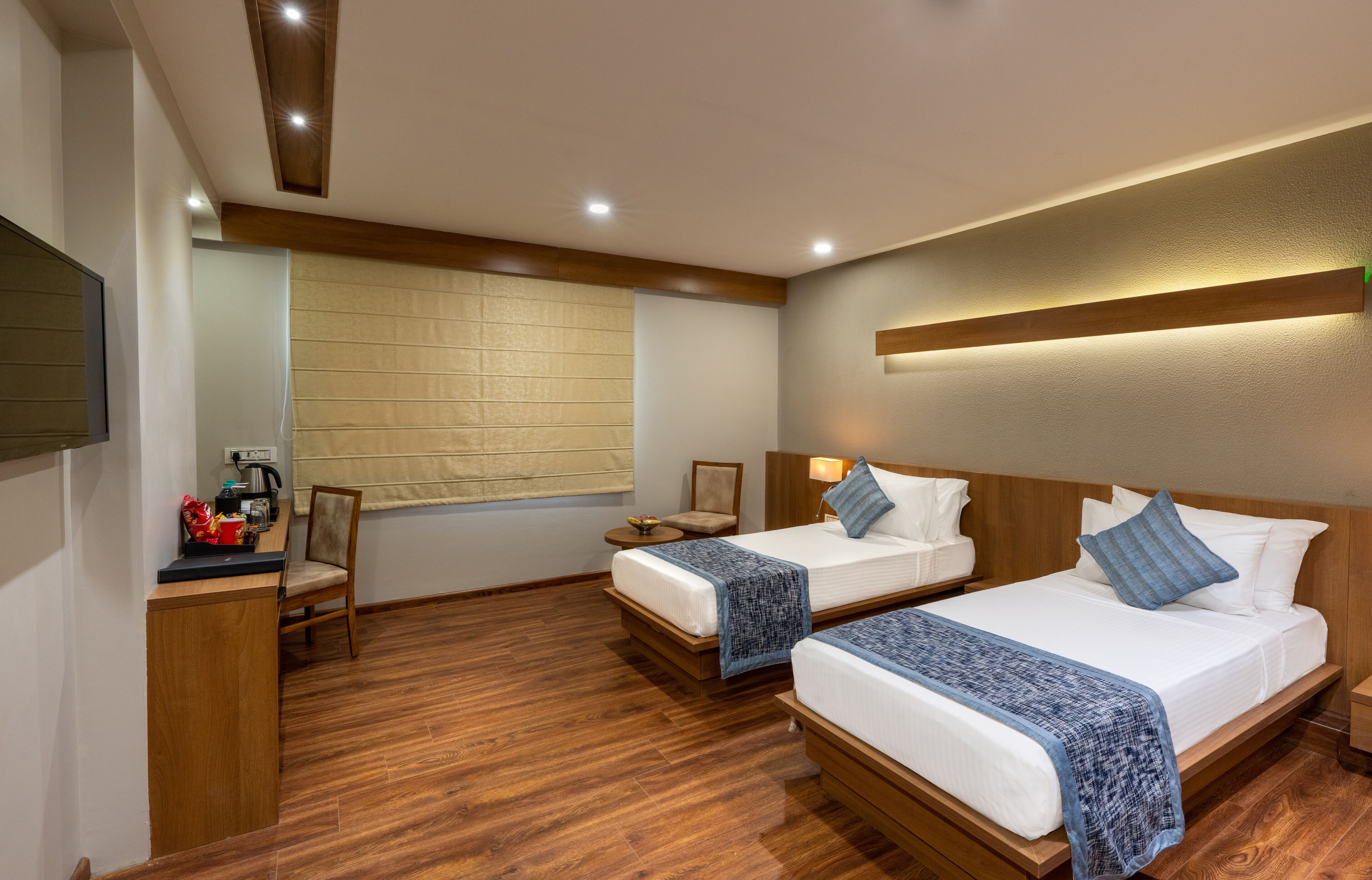 Habitación de hotel con dos camas, suelo de madera, un escritorio con silla, televisión y ventana con persianas.