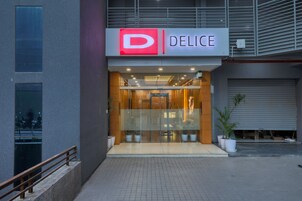 Un edificio moderno con un letrero que dice 'D' y 'DELICE', ventanas amplias y plantas en macetas afuera.