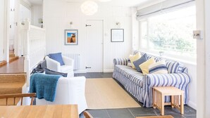 A bright, spacious living area with a white sofa, a striped sofa, and a dining table. There is a door in the background and a window providing natural light.